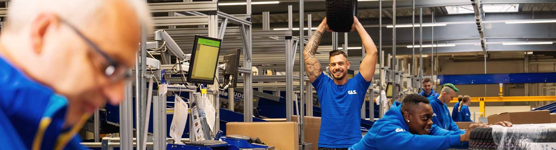 Employees are working together in the warehouse; one colleague is holding up a car tire