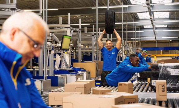Employees are working together in the warehouse; one colleague is holding up a car tire