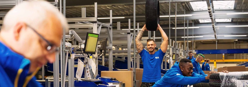 Employees are working together in the warehouse; one colleague is holding up a car tire