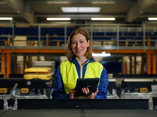 A GLS employee is standing by the conveyor belt with a tablet and a safety vest