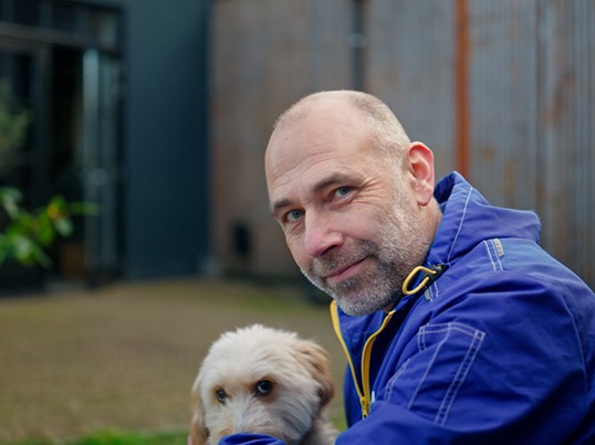 A GLS employee in work clothes looks at the camera, and a small dog is sitting next to him